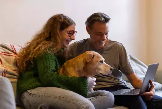 a man and woman sit on the sofa smiling with their dog talking into a laptop during a telemedicine visit