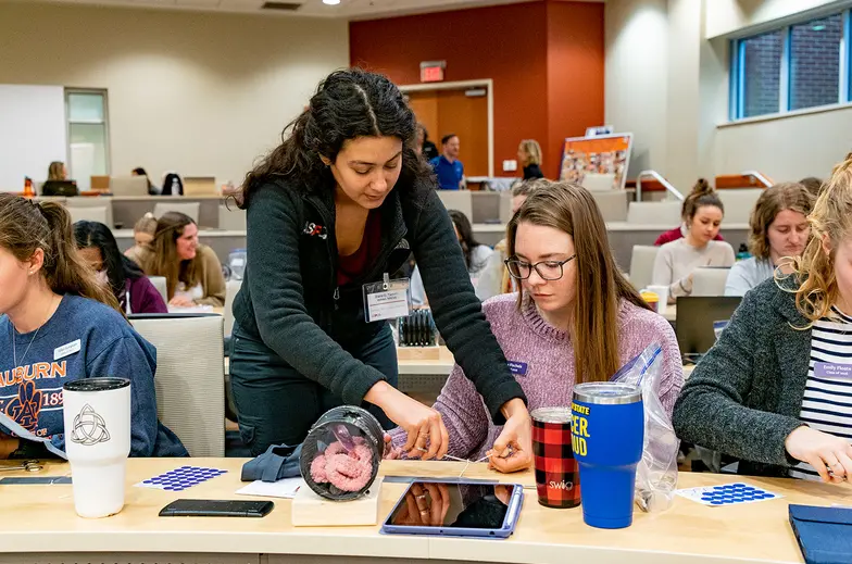 two veterinary students work together at a desk in a room full of trainees, one student stands to assist the other 