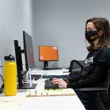 Person wearing ASPCA branded mask and jacket sitting at desk in front of computer monitor