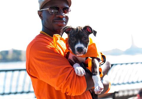 person outdoors in orange tee shirt holding tan and black puppy in an orange bandanna