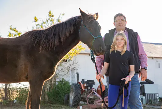 a young blonde person stands with an adult holding the reins of a brown horse outdoors near a barn structure