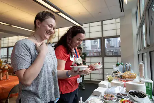 two women smile while creating sundaes at an office party buffet