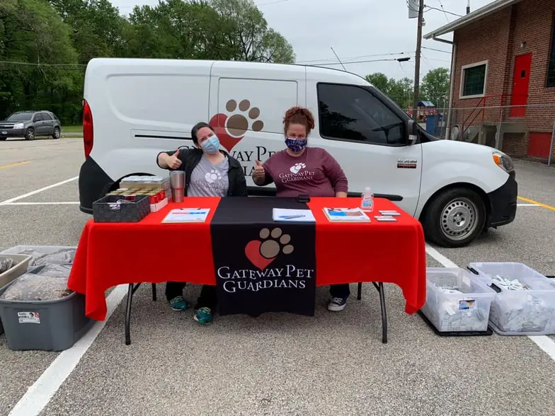 two workers sit at a table in front of a pet food distribution van giving the thumbs up sign