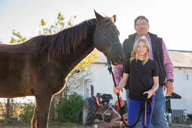 a young blonde person stands with an adult holding the reins of a brown horse outdoors near a barn structure