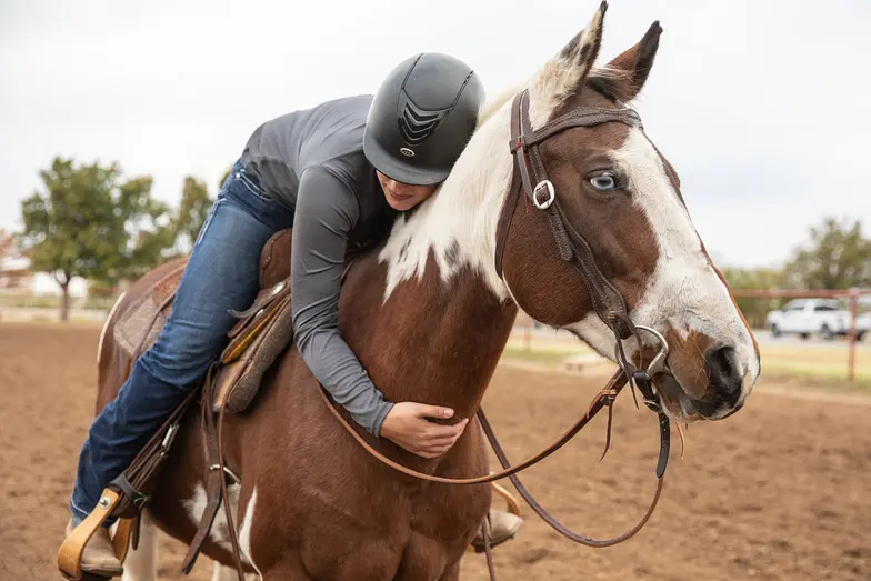 a rider in helmet and jeans leans over atop brown and white horse to hug him