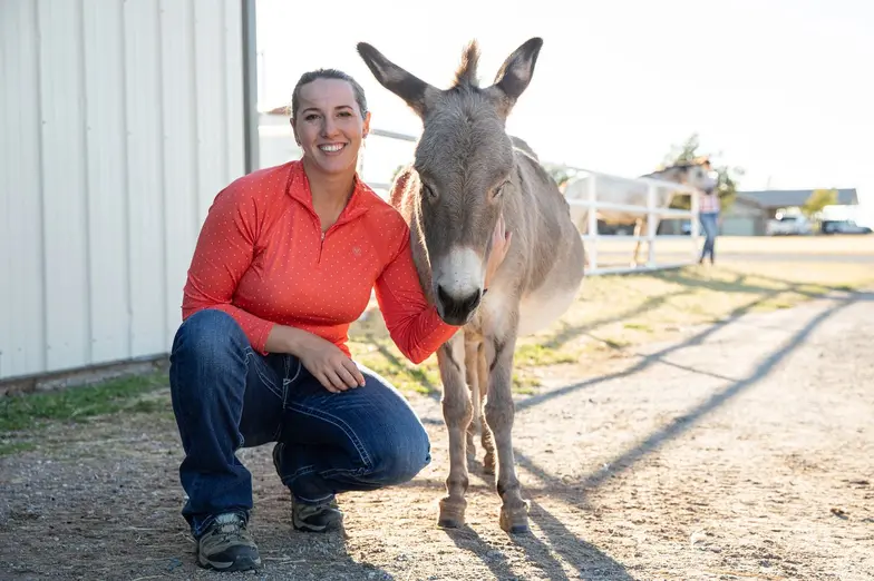 a woman wearing an orange top kneels next to a tan donkey near a barn structure